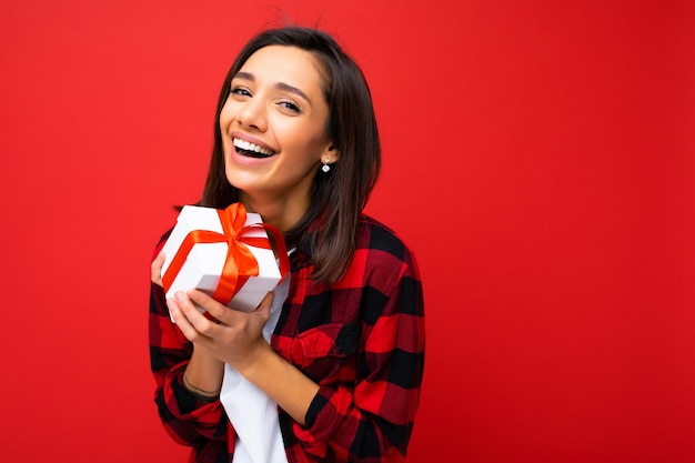 attractive positive smiling young brunette woman isolated over colourful wall wearing everyday trendy outfit holding gift box