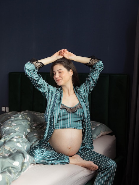 Attractive pregnant woman in pajamas sits in bed and holds her belly Last months of pregnancy