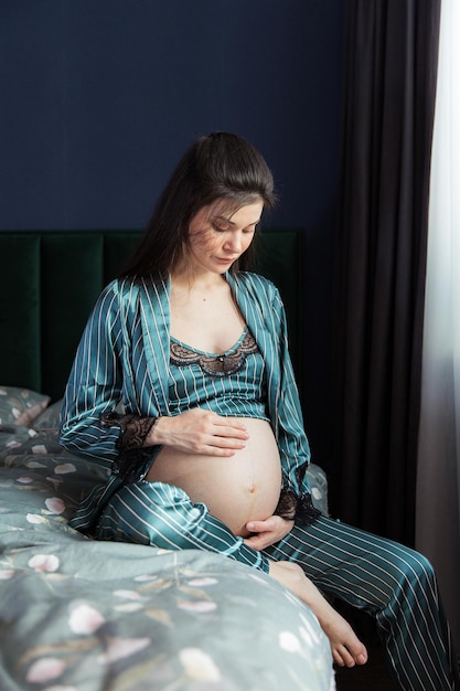 Attractive pregnant woman in pajamas sits in bed and holds her belly Last months of pregnancy