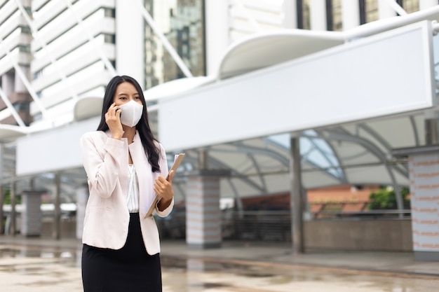 Attractive professional asian business woman using mobile phone wearing protective face mask prevent covid-19 virus  in the city outdoor