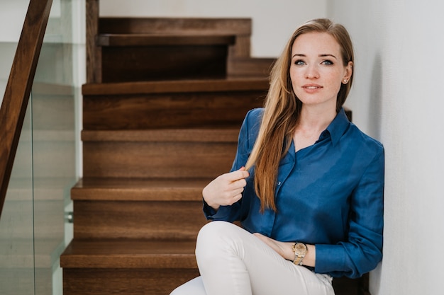 Attractive red haired young woman dressed in a blue shirt and white pants sitting on stairs at her house, touching hair, looks confident and satisfied by her life. stay home. Happy people concept.