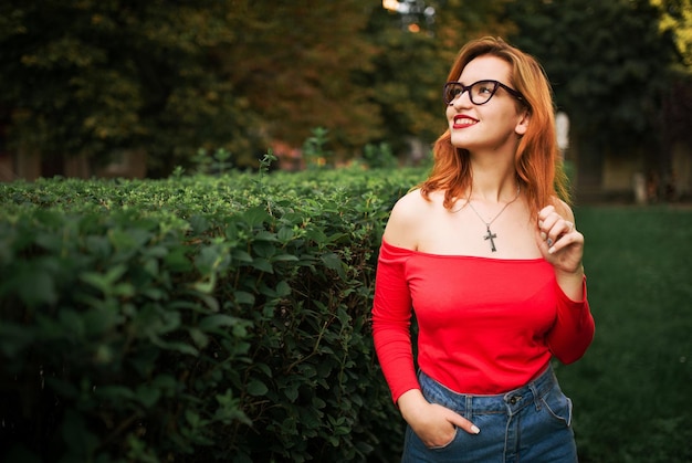 Attractive redhaired woman in eyeglasses wear on red blouse and jeans skirt posing at green park