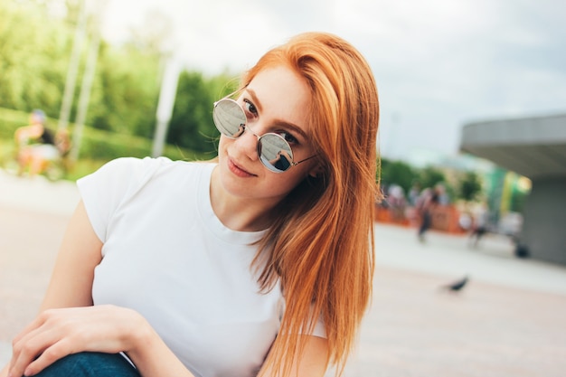 Attractive redhead smiling girl in round sunglasses in casual clothes sitting on street in city