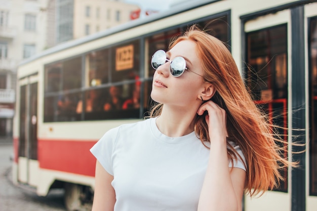 Attractive redhead smiling girl in sunglasses in casual clothes on tram background at street