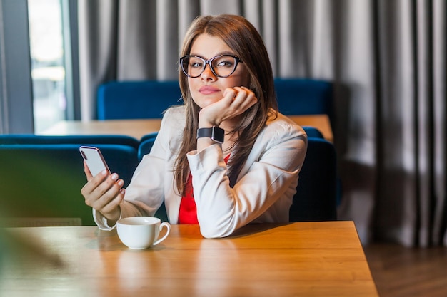 Attractive relaxed brunette young woman in glasses sitting holding cup of coffee drink and smart phone in hands, looking at camera with calm expression. Indoor shot, cafe or office background.