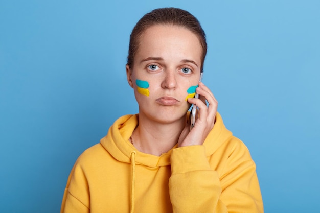 Attractive sad upset woman in hoodie with Ukrainian flag on cheeks posing isolated over blue background talking phone hearing bad news about situation in Ukraine stop war