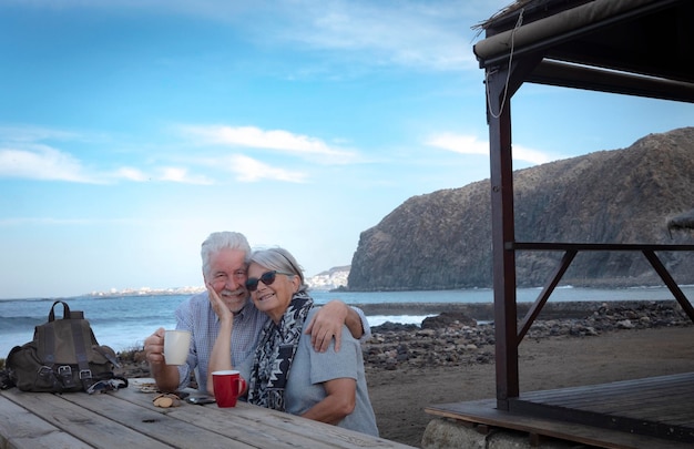 Attractive senior couple exchanging a tender gesture on the beach Sitting at a wooden table with two cups of coffee Horizon over the sea