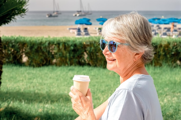 Attractive senior woman grayhaired wearing blue sunglasses sitting face to the beach holding a takeaway coffee enjoying a sunny day