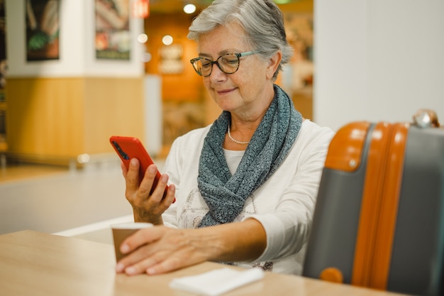 Attractive senior woman sitting at the airport cafe using social media on mobile phone while waiting for boarding. Happy mature traveler with luggages