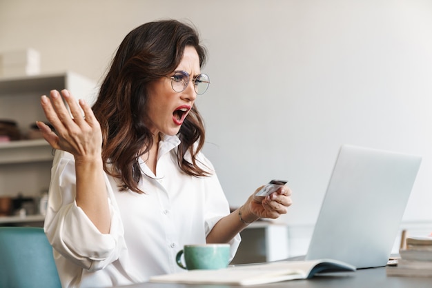 Attractive shocked upset young brunette businesswoman sitting at the cafe table with laptop computer indoors, shopping online with credit card