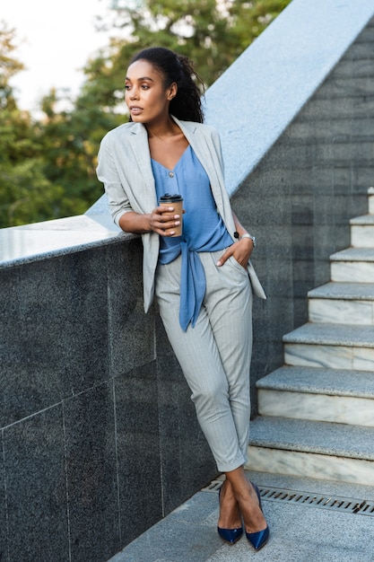 Attractive smiling african business woman standing outdoors at the city, leaning on stairs, holding takeaway coffee cup