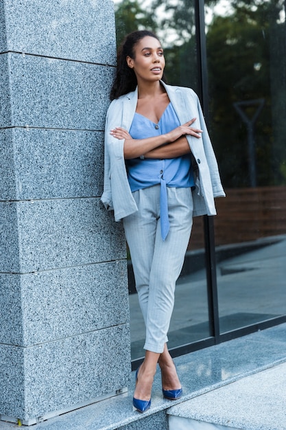 Attractive smiling african business woman standing outdoors at the city, leaning on a wall