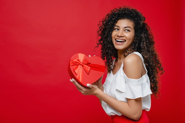 Attractive smiling black woman holding heart shape gift isolated over red