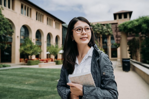 Attractive smiling charming asian young girl student with school book relaxing walking in university outdoor. college lady enjoy sunshine at the park in stanford surrounding by green grass and trees.