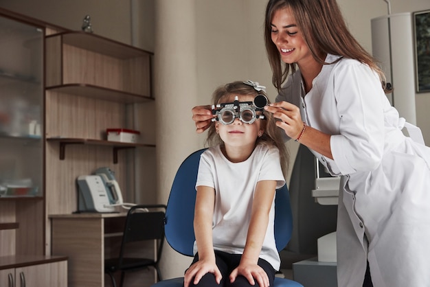 Attractive smiling female doctor loving her job. Little girl in glasses sitting in clinic and having her eyes tested.