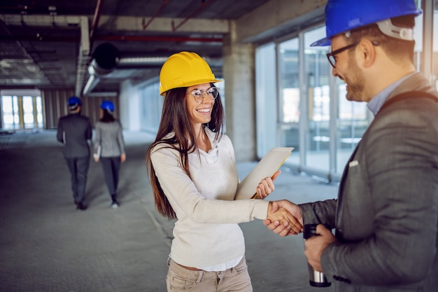 Attractive smiling friendly caucasian female architect shaking hands with her colleague and congratulations him on great idea for building renewal.