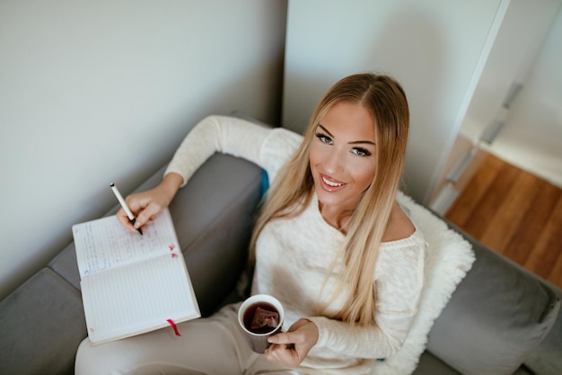 Attractive smiling girl sitting at her living room with cup of tea and writing something at the notebook. Looking at camera. Top view.