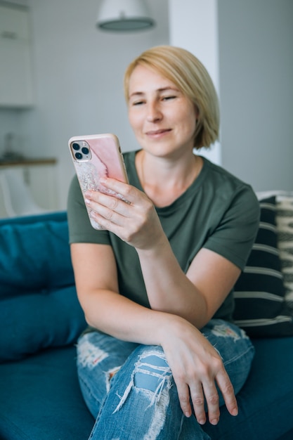 Attractive smiling middle age senior woman using smart phone while sitting on the sofa at home