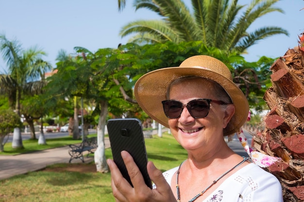 Attractive smiling retired woman in a public park using smart phone to talk with friends active lifestyle for a pensioner people in nature under the sun