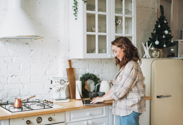 Attractive smiling woman curly hair in plaid shirt using laptop at bright kitchen