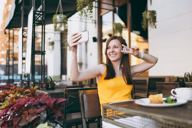 Attractive smiling woman in outdoors street coffee shop cafe sitting at table, listen music in headphones, doing selfie shot on mobile phone, relaxing in restaurant free time