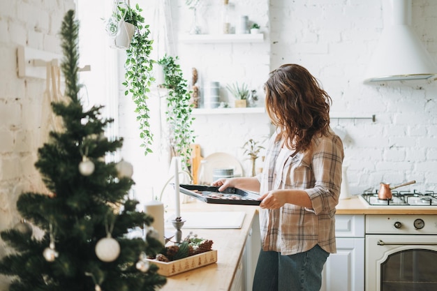 Attractive smiling woman with curly hair in plaid shirt bakes cookies at bright kitchen at the home