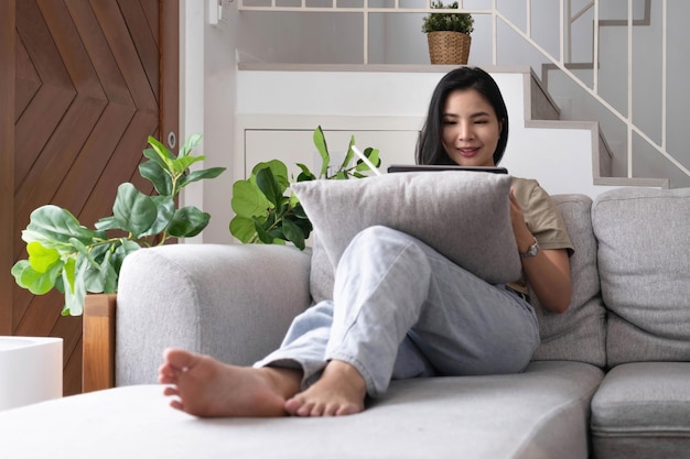 Attractive smiling young asian woman relaxing on a leather couch at home working on laptop computer