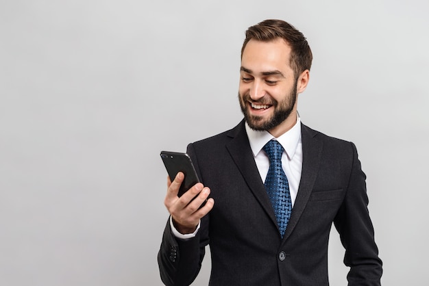 Attractive smiling young businessman wearing suit standing isolated over gray wall, holding mobile phone