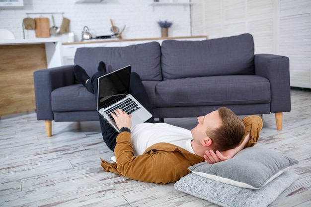 Attractive smiling young man in casual wear sitting on floor on sofa in living room using laptop computer