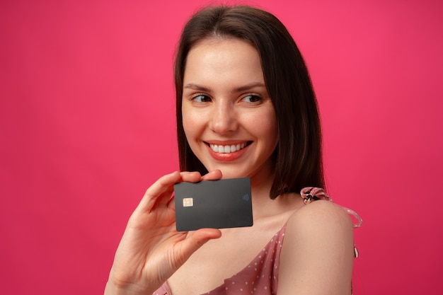 Attractive smiling young woman holding black credit card against pink studio surface