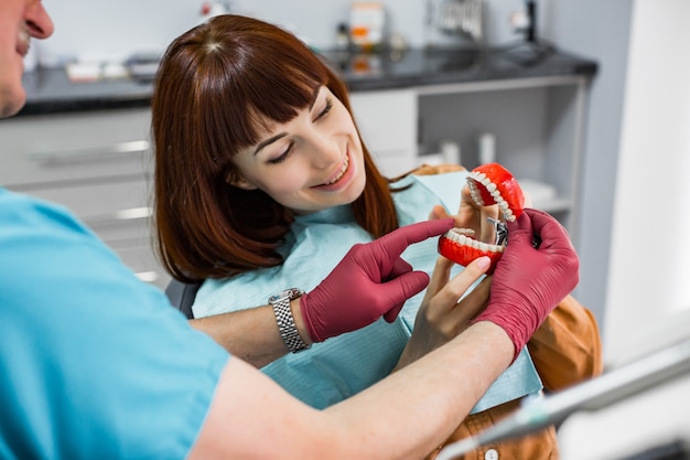 Attractive smiling young woman sitting in dental chair in stomatology clinic with senior male dentist, showing her jaw and teeth sample. Healthy teeth concept.