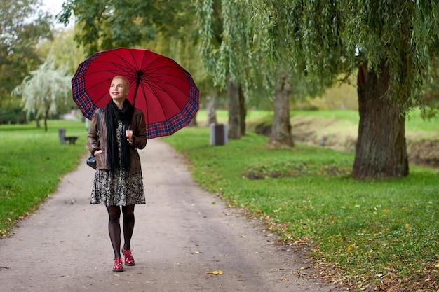 Attractive smilling short haired blonde woman walking in the autumn park with huge red umbrella