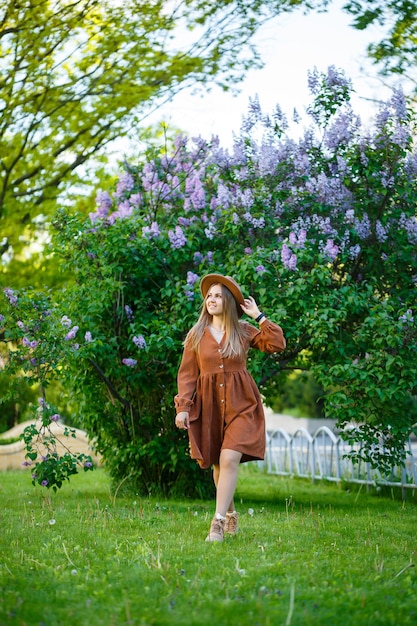 Attractive stylish smiling slim girl with long hair walks in the park, dressed in a warm brown dress, autumn fashionable street fashion, with a hat. Young woman of European appearance