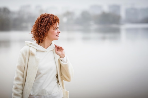 Attractive stylish woman dressed in white warm coat holding yellow leaves walking near river