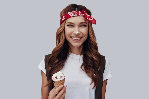 Attractive stylish young woman eating ice cream and smiling while standing against grey background