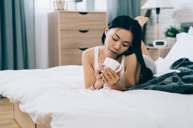 attractive taiwanese woman lying on belly in bedroom is looking into the cup. seductive asian female wearing skimpy clothes is having coffee on white bedding.