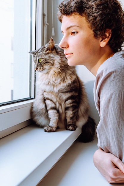 Attractive teenage girl next to beloved cat hugging while looking out window