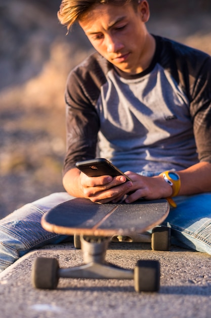 Attractive and thoughtful teenager boy relaxing with a skateboard and sitting down on a bench holding and using a smartphone for networking during a sunny day, outdoors