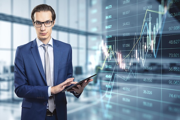 Attractive thoughtful young european businessman with tablet and glowing forex index chartgraph standing in blurry office interior Trade finance and market concept Double exposure