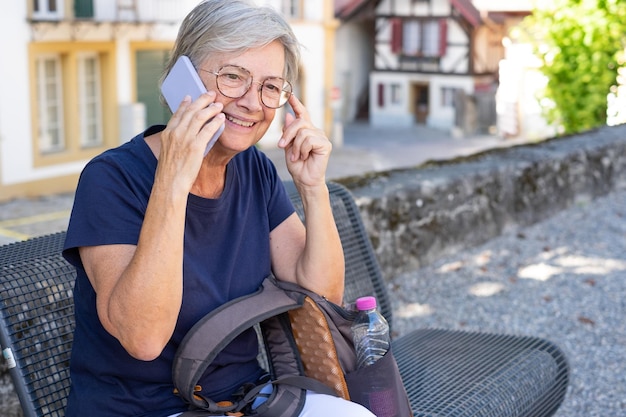 Attractive traveler senior woman sitting on a bench visiting an old village talking at mobile phone