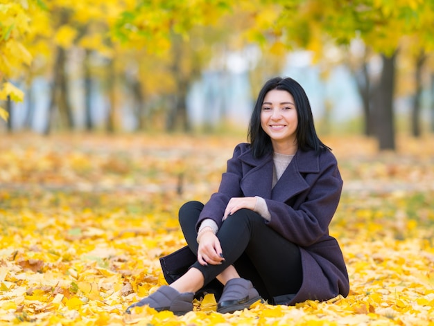 Attractive trendy young woman sitting amongst autumn leaves in a park smiling happily at the camera