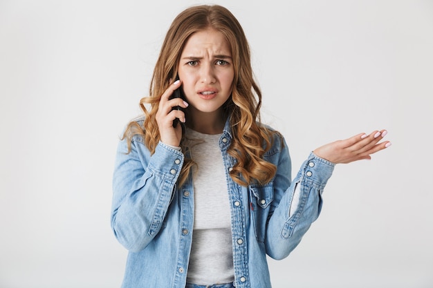 Attractive upset young girl standing isolated over white , talking on mobile phone