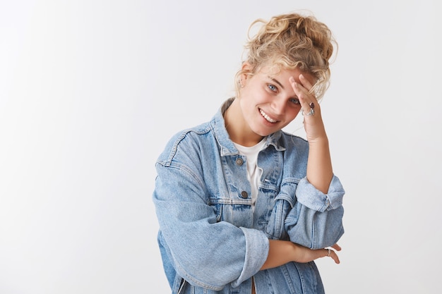 Attractive urban curly-haired blond caucasian woman curly stylish hairstyle touching forehead smiling tilting head carefree enjoying party, talking laughing pleased, standing white wall