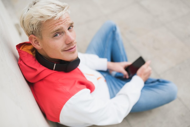 Attractive above view of young blond man looking up and enjoying weather in city park waiting for his friends texting message with headphones on the neck outside in the park