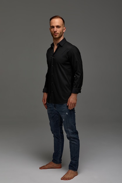 An attractive white man in jeans and black shirts tands in full growth against a gray background Copy space