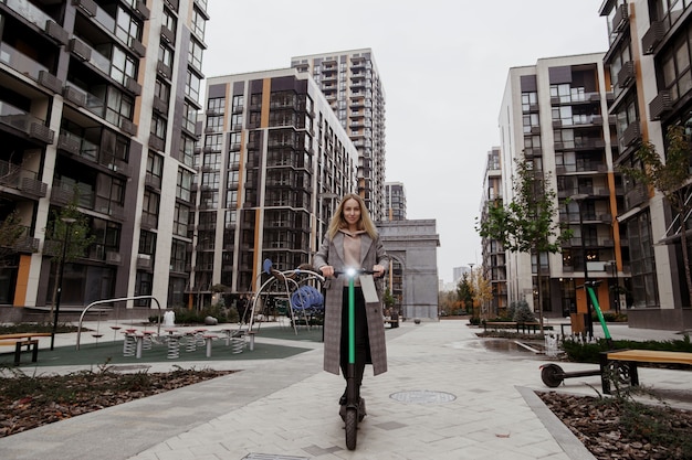 Attractive woman in casual clothes rides rented electric scooter. Apartment blocks on background. Comfort way to trip around the city. Fast trip concept. Eco habits.