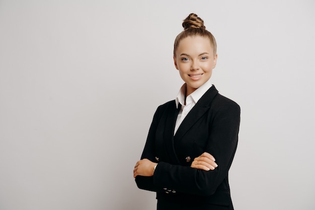 Attractive woman executive in dark suit with hair in bun, standing sideways looking forward with her arms crossed radiating calmness and confidence isolated on grey background. Business people concept