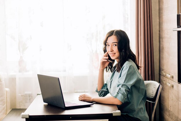 Attractive woman having happy look while speaking over smartphone, using black laptop. People, technology, lifestyle, working at home. Business woman in casual clothes consulting customer on phone