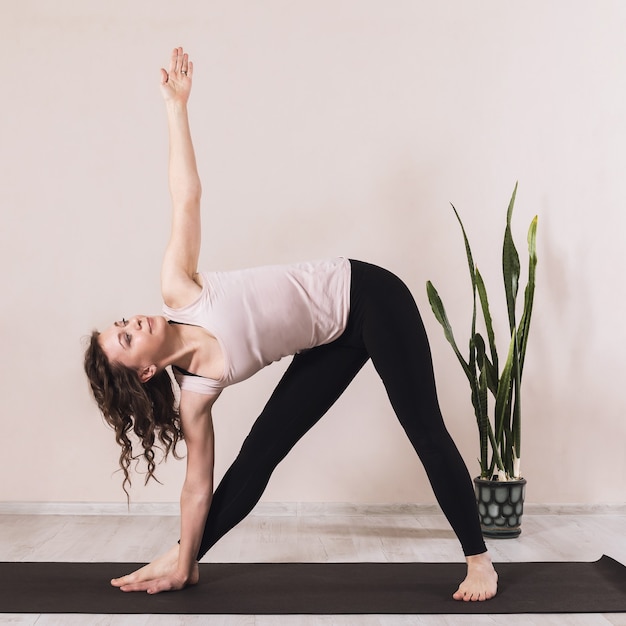 Attractive woman is engaged in yoga performs the exercise Utthita Trikonasana triangle pose trains in sportswear standing on a mat near the wall