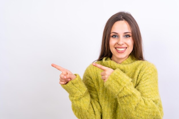 Attractive woman smiling pointing fingers at copy space on white background green wool sweater